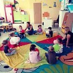 Dzieci z wychowawczynią siedzą na wielokolorowej, okrągłej płachcie