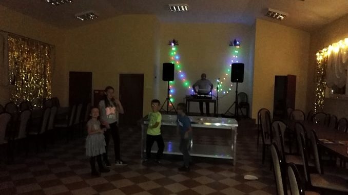 Wnetrze sali w Domu Ludowym w Woli Rokszyckiej - czworo dzieci stoi przy nowym sprzęcie nagłaśniającym; za mimi na poswyższonej scenie mężczyzna przy stoje didżejskim