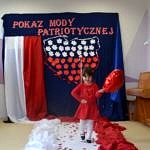 Przedszkolaki prezentują sie w biało-czerwonych strojach. Na ścianie na granatowym tle napis "Pokaz mody patriotycznej", poniżej kształt Polski wypełniony biało-czerwonymi liśćmi, obok biało-czerwona flaga. Przedszkolaki chodzą po biało-czewonym podeście - dywanie