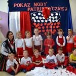Przedszkolaki prezentują się w biało-czerwonych strojach. Na ścianie na granatowym tle napis "Pokaz mody patriotycznej", poniżej kształt Polski wypełniony biało-czerwonymi liśćmi, obok biało-czerwona flaga. Grupa przedszkolna z opiekunką w ludowym stroju
