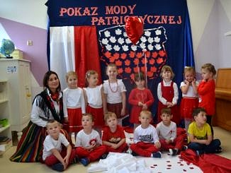 Przedszkolaki prezentują się w biało-czerwonych strojach. Na ścianie na granatowym tle napis "Pokaz mody patriotycznej", poniżej kształt Polski wypełniony biało-czerwonymi liśćmi, obok biało-czerwona flaga. Grupa przedszkolna z opiekunką w ludowym stroju