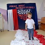 Przedszkolaki prezentują sie w biało-czerwonych strojach. Na ścianie na granatowym tle napis "Pokaz mody patriotycznej", poniżej kształt Polski wypełniony biało-czerwonymi liśćmi, obok biało-czerwona flaga. Przedszkolaki chodzą po biało-czewonym podeście - dywanie