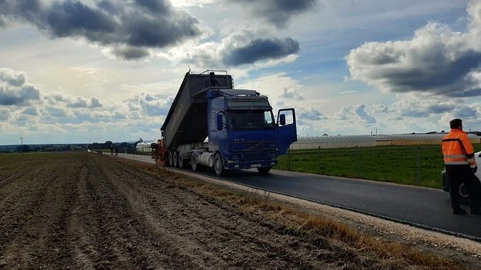 Nowa droga asfaltowa, obok zaorane pole, na niej samochód ciężarowy wykonujący prace drogowe wszystko na tle pokrytego chmurami nieba