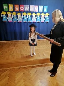 Dziewczynka w stroju galowym i birecie pasowana na "zerówkowicza" przez dyrektor szkołay Adriannę Grelę
