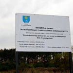 Tablica informacyjna z herbem gminy informująca o dofinansowaniu z Funduszu Dróg Samorządowych przebudowy drogi "bez nazwy" w Woli Krzysztoporskiej