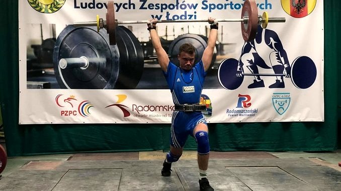 Zawodnik w niebieskiej koszulce podnosi sztanę; w tle baner z napisem Wojewódzki Turniej o Puchar Przewodniczącego LZS w Łodzi