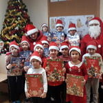 Grupa dzieci w mikołakowych czerwono-białych czapeczkach stoi przy choince; dzieci trzymaja w dłoniach kalendarze adwentowe; obok stoi Mikołaj