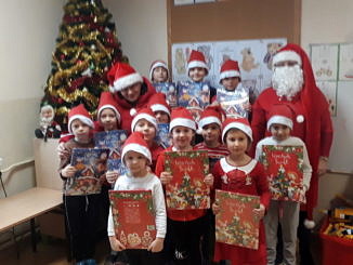 Grupa dzieci w mikołakowych czerwono-białych czapeczkach stoi przy choince; dzieci trzymaja w dłoniach kalendarze adwentowe; obok stoi Mikołaj