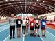 Grupa młodych lekkoatletów z numerami sartowymi na piersiach stoi z trenerem na dużej hali sportowej o półokrągłym sklepieniuz trenerem