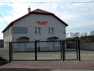 Widok Domu Ludowego w Kacprowie, a przed nim utwardzony kostką betonową teren i nowe ogrodzenie panelowe z bramą i furtką