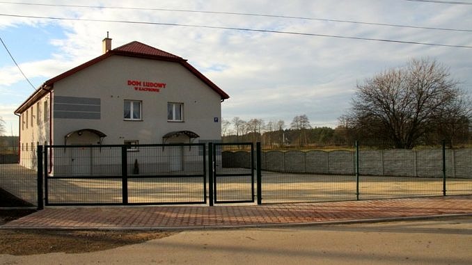 Widok Domu Ludowego w Kacprowie, a przed nim utwardzony kostką betonową teren i nowe ogrodzenie panelowe z bramą i furtką