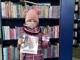 Dziewczynka z książką i kartą biblioteczną stoi na tle regałów z ksiązkami