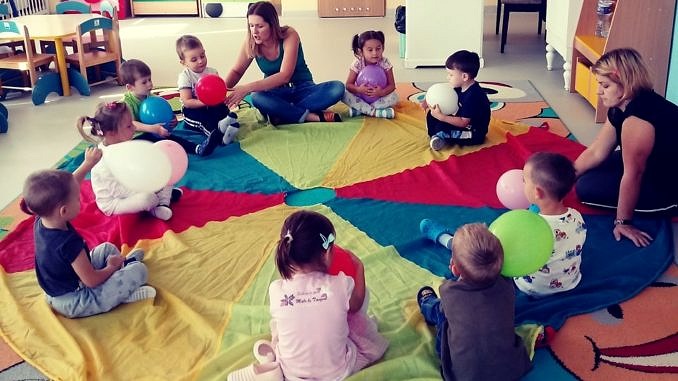 Dzieci z opiekunkami siedzą w kręgu na kolorowej tęczowej macie i podają sobie balonik
