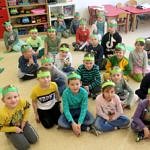 Grupa dzieci przedszkolnych siedzi na podłodze z zielonymi opaskami z dinozaurami na głowach