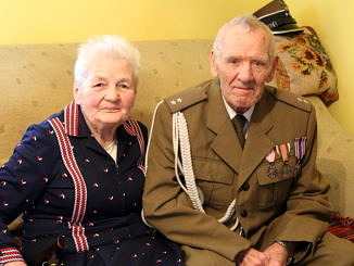 Dwoje starszych ludzi siedzi na wersalce - pani Zofia w granatowej sukience w czerwone paski; obok pan Zdzisław w mundurze wojskowym