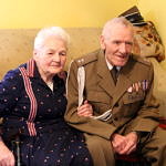 Dwoje starszych ludzi siedzi na wersalce - pani Zofia w granatowej sukience w czerwone paski; obok pan Zdzisław w mundurze wojskowym