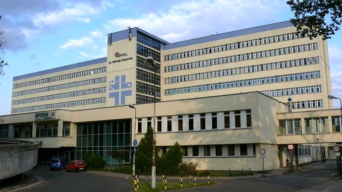 Front szpitala im. Kpernika w Łodzi; szary, duży budynek; nad wejściem logo w postaci niebieskiego podwójnego krzyża