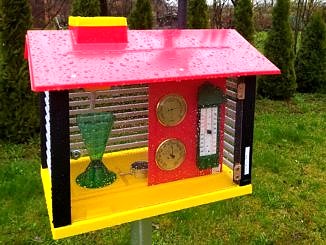 Stacja meteorologiczna - kolorowa skrzynka ze spadzistym czerwonym daszkiem i żółtą podłogą, a w środku urzadzenia do pomiaru pogody; nad daszkiem zamontowany Kur (kogut) do pomiaru kierunku wiatru