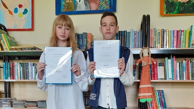 Dwoje młodych ludzi - dziewczyna i chłopak - stoją na tle regalu z książkami i trzymaja w dłoniach zaświadczenia, że są finalistami konkursu przedmiotowego