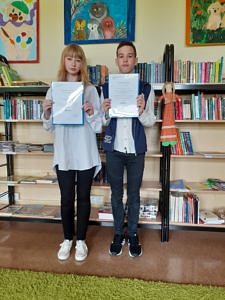 Dwoje młodych ludzi - dziewczyna i chłopak - stoją na tle regalu z książkami i trzymaja w dłoniach zaświadczenia, że są finalistami konkursu przedmiotowego