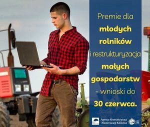 Młody mężczyzna w kraciastej koszuli trzyma laptopa; wszystko na tle pola i ciągnika