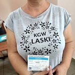 Kobieta w koszulce z napisem KGW Laski trzyma w dłoniach certyfikat potwierdzjący szczepienie