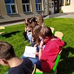 Dzieci siedzą w kręgu przed szkołą na krzesłach ustawionych na trawie - obok tablica z hasłami Narodowego Czytania