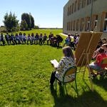 Dzieci siedzą w kręgu przed szkołą na krzesłach ustawionych na trawie - obok tablica z hasłami Narodowego Czytania