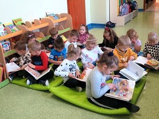 Dzieci siedzą na macie przy regale z ksiązkami i przeglądają książki