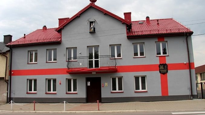 Dom Ludowy w Woli Krzysztoporskiej - szary budynek z czerwonym dachem i czerwonymi ozdobnymi pasami. na elewacji znak starży pożarnej