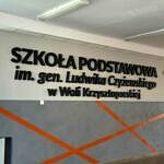 szary korytarz szkolny z czarnym napisem Szkoła Podstawowa im. gen. Ludwika Czyżewskiego w Woli Krzysztoporskiej