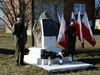 Apel Pamięci - chłopcy w mundurach stoja przy obelisku w Bujnach w tle flagi